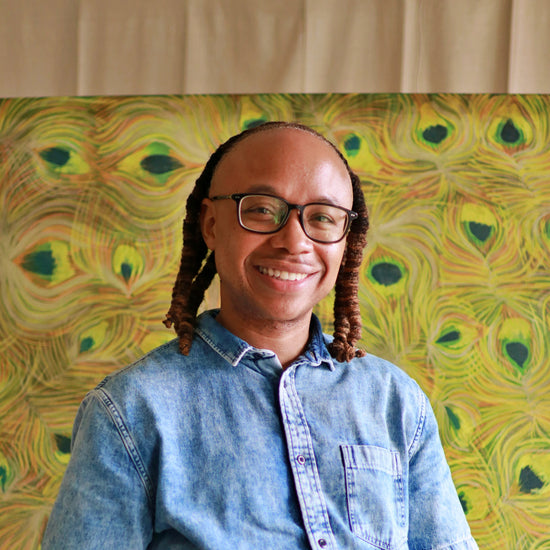 Person wearing glasses and a blue shirt sitting in front of a peacock feather patterned wall.