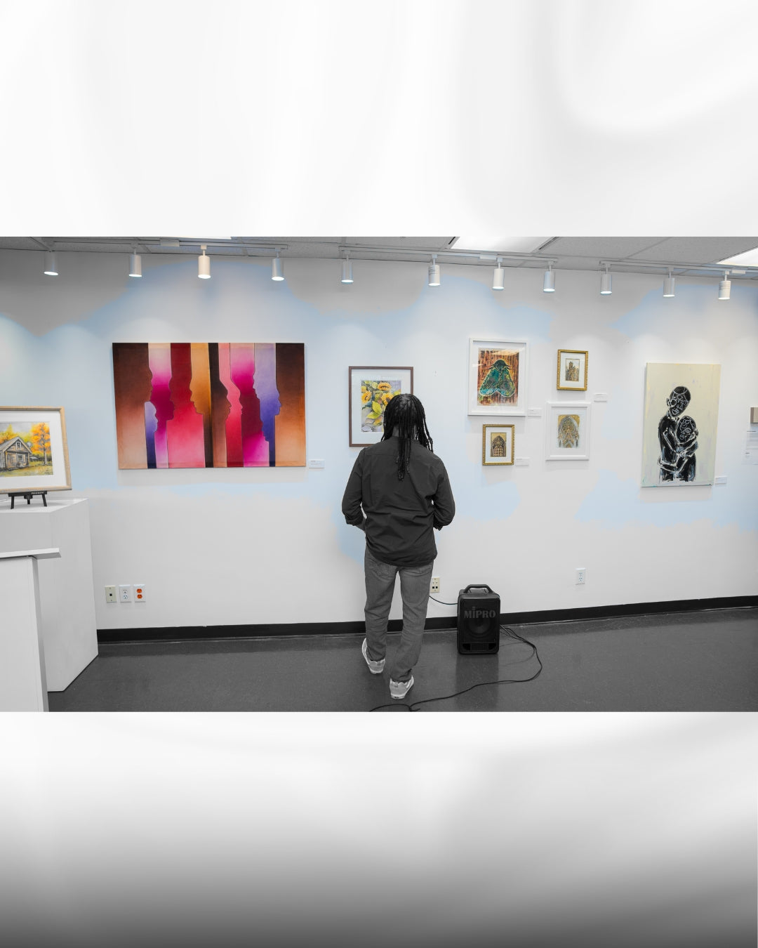 A person stands with their back to the camera in an art gallery, looking at a colorful abstract painting mounted on a white wall. Several other framed artworks are displayed on the gallery wall under track lighting.